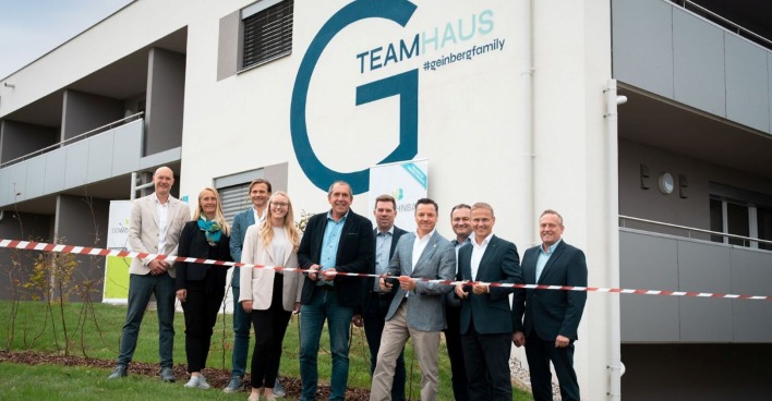 Zu sehen ist das neue Teamhaus für die Mitarbeiter des Spa Resort Geinberg. Im Vordergrund eine Gruppe von Menschen, die sich über die Eröffnung freuen. Einer der Anwesenden durschneidet ein rotes Band.