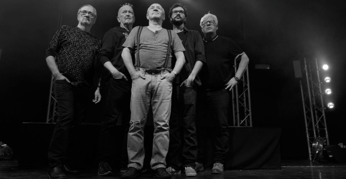 Schwarz-Weiß-Foto einer fünfköpfigen Band auf der Bühne, die nebeneinander in die Kamera blickt. Die Musiker tragen legere Kleidung und stehen vor einer Bühnenkulisse mit Scheinwerfern.