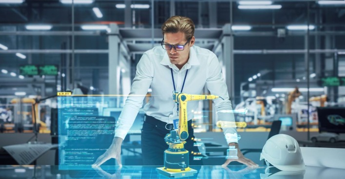 Ein Ingenieur in einem modernen Industriegebäude steht über einem Tisch und betrachtet ein holografisches 3D-Modell eines gelben Roboterarms mit eingeblendeten Programmiercodes und technischen Daten. Neben ihm liegt ein weißer Schutzhelm auf dem Tisch.