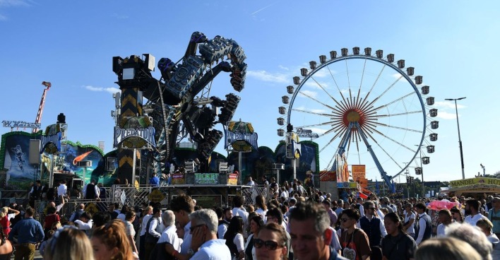 Menschenmenge auf dem Oktoberfest in München, im Hintergrund ein großes Riesenrad und Fahrgeschäfte bei blauem Himmel. Leiche hinter dem Riesenrad auf dem Oktoberfest in München entdeckt – Polizei vermutet medizinischen Notfall.