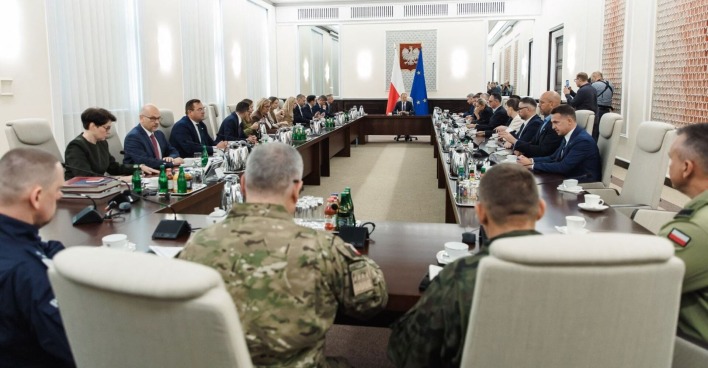 Langer Konferenztisch mit polnischen Regierungsmitgliedern und Militärs in Uniform, Donald Tusk sitzt am Kopfende zwischen polnischer und EU-Flagge.