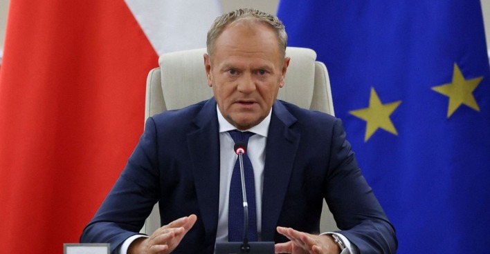 Donald Tusk sitzt in einem dunklen Anzug vor Mikrofonen zwischen einer polnischen und einer EU-Flagge bei einer Krisensitzung.