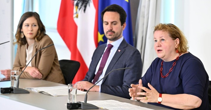 Drei Personen sitzen bei einer Pressekonferenz an einem Tisch mit Mikrofonen und Unterlagen, im Hintergrund sind österreichische und EU-Flaggen zu sehen.