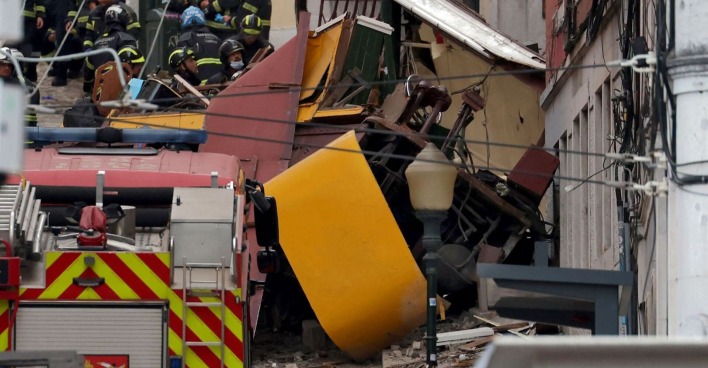 Zerstörter gelber Waggon der Standseilbahn „Elevador da Glória“ nach dem schweren Unfall in Lissabon, umgeben von Feuerwehrleuten und Rettungskräften.