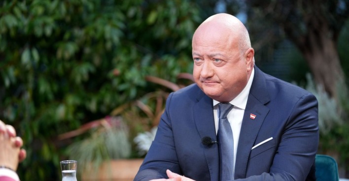 Christian Stocker im dunklen Anzug mit Krawatte und Österreich-Anstecker sitzt bei einem Fernsehinterview im Freien. Vor ihm steht ein Glas Wasser, im Hintergrund sind Pflanzen zu sehen.