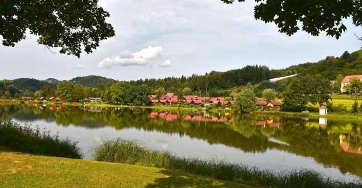 Blick über den Sulmsee in der Steiermark, mit spiegelglattem Wasser, roten Hütten am Ufer und dicht bewaldeten Hügeln im Hintergrund.