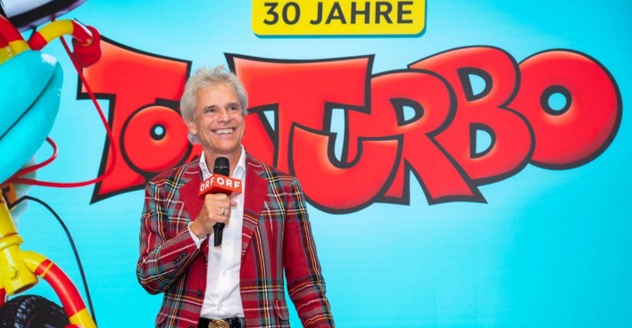 Thomas Brezina hält ein ORF-Mikrofon, lächelt vor einer großen „Tom Turbo“-Jubiläumswand mit der Aufschrift „30 Jahre“.