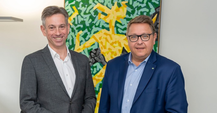 Werner Ressi und Martin Graf stehen nebeneinander vor einem farbenfrohen abstrakten Gemälde und lächeln in die Kamera.