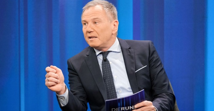 Armin Wolf sitzt in einem TV-Studio vor blauem Hintergrund und gestikuliert während eines Gesprächs.