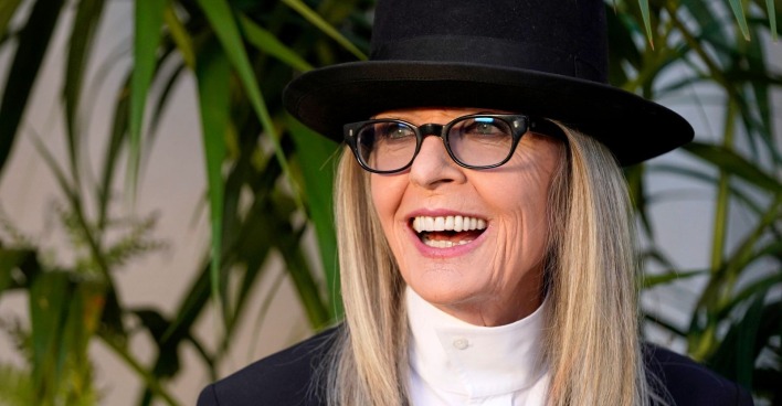 Diane Keaton, lachend, trägt einen schwarzen Hut und eine Brille; sie steht vor grünen Pflanzen.