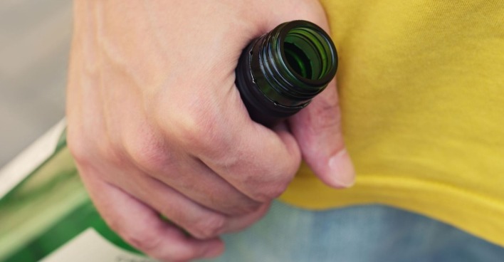 Mann mit gelbem T-Shit hält in aggressiver Grundhaltung Weinflasche aus Grünglas in seiner rechten Hand