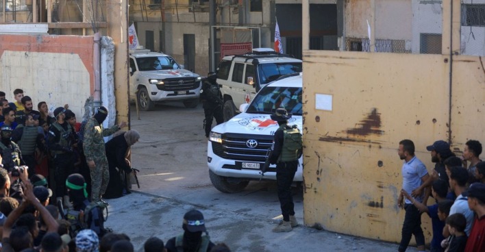 Weiße Fahrzeuge des Roten Kreuzes fahren durch ein Tor im Gazastreifen, bewaffnete Kämpfer sichern die Szene, während Menschen am Straßenrand zusehen.