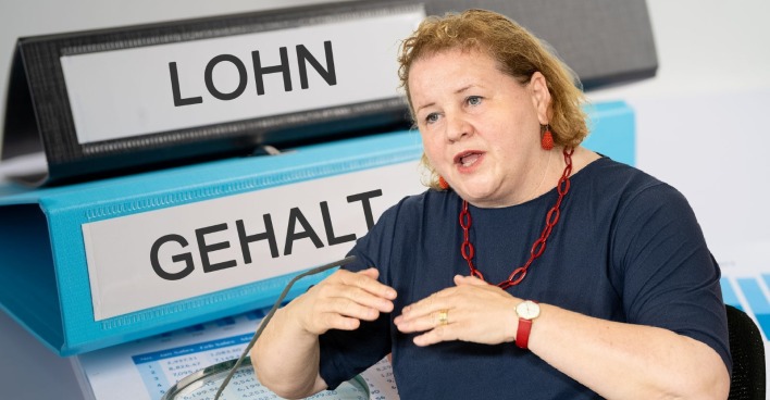 Korinna Schumann spricht über das Lohntransparenz-Gesetz, im Hintergrund Aktenordner mit der Aufschrift „Lohn“ und „Gehalt“.