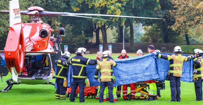 Feuerwehrleute und Sanitäter in Herdecke schirmen eine verletzte Person beim Transport zu einem roten Rettungshubschrauber ab.