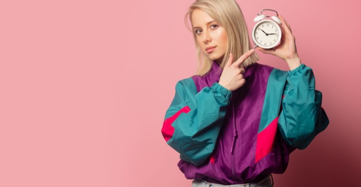 junge blonde Frau in einem bunten Blouson und mit einem klassischen Wecker in der Hand