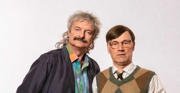 Zwei Männer in typischer 90er-Jahre-Aufmachung stehen nebeneinander vor hellem Hintergrund – das legendäre Comedy-Duo „Weber &amp; Breitfuß“ aus der ORF-Kultserie.