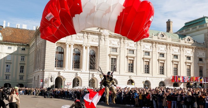Ein Fallschirmspringer des österreichischen Bundesheeres mit rot-weißem Schirm landet am Heldenplatz in Wien vor einer jubelnden Menschenmenge während der Feierlichkeiten zum Nationalfeiertag.