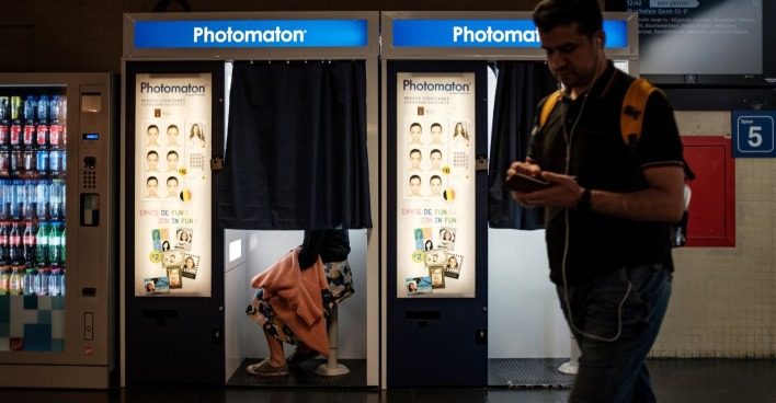 Fotobox im Bahnhofsgebäude, teilweise geschlossener Vorhang, eine Person sitzt drinnen, daneben ein Passant mit Rucksack – Symbolbild für den Vorfall am Rostocker Hauptbahnhof.