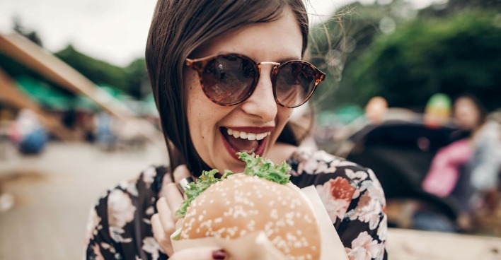 Frau mit Sonnenbrille hält lächelnd einen Burger auf einem Street-Food-Festival in der Hand.