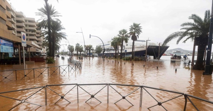 Überflutete Straßen im Hafen von Ibiza Stadt mit stehenden Wassermassen, Palmen und festliegenden Fähren im Hintergrund.