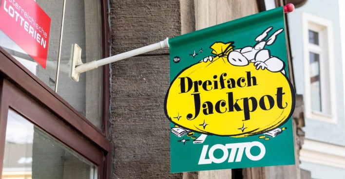 Werbeschild der Österreichischen Lotterien mit der Aufschrift „Dreifach Jackpot“ vor einer Lotto-Annahmestelle.