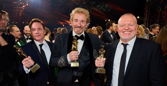 Thomas Gottschalk hält bei der Kurier Romy 2025 in Kitzbühel seine Diamant-Romy in den Händen, flankiert von den zwei weiteren Sonderpreisträgern Bully Herbig und Stefan Raab, umgeben von Gästen in festlicher Abendgarderobe.