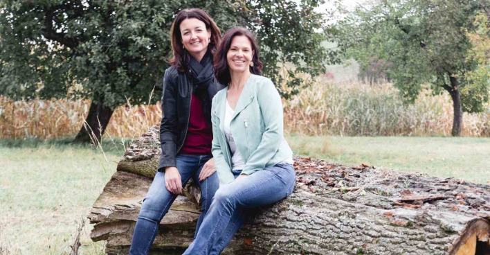 Zwei lächelnde Frauen sitzen auf einem umgestürzten Baumstamm in einer herbstlichen Wiese mit Bäumen und Maisfeld im Hintergrund.
