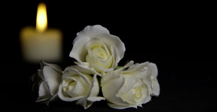 Schöne weiße Rosen mit einer brennenden Kerze auf dem dunklen Hintergrund. Trauerblume und Kerze auf dem Tisch vor schwarzem Hintergrund mit Kopierraum.