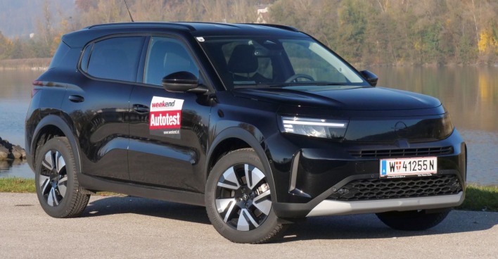 Ein schwarzer Opel Frontera parkt am Rande eine Gewässers. Das Auto leuchtet in der Sonne, im Hintergrund sind einige Bäume zu erkennen.
