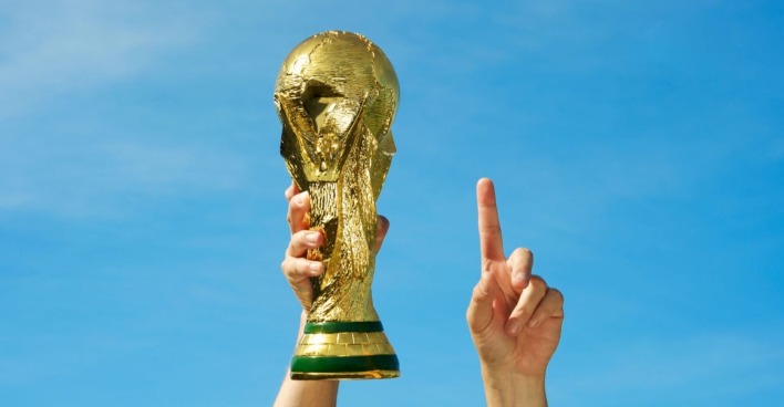 Mensch, der in seiner linken Hand WM-Pokal hält und seinen rechten Zeigefinger nach oben streckt