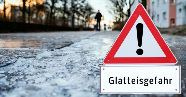 Verkehrswarnschild auf vereister Straße mit deutlicher Glatteisgefahr; glatte Oberfläche und Eisplatten zeigen das erhöhte Risiko durch aktuelle Glatteiswarnung.
