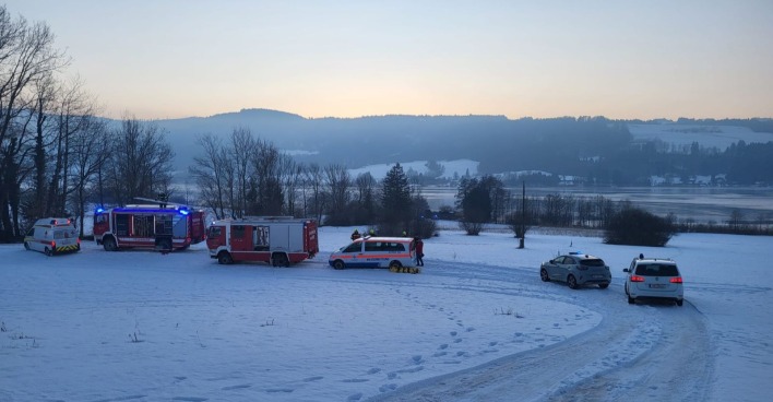 Mehrere Einsatzfahrzeuge von Feuerwehr, Wasserrettung und Polizei stehen auf einer verschneiten Wiese am Irrsee in Oberösterreich während eines Rettungseinsatzes bei winterlicher Abenddämmerung.