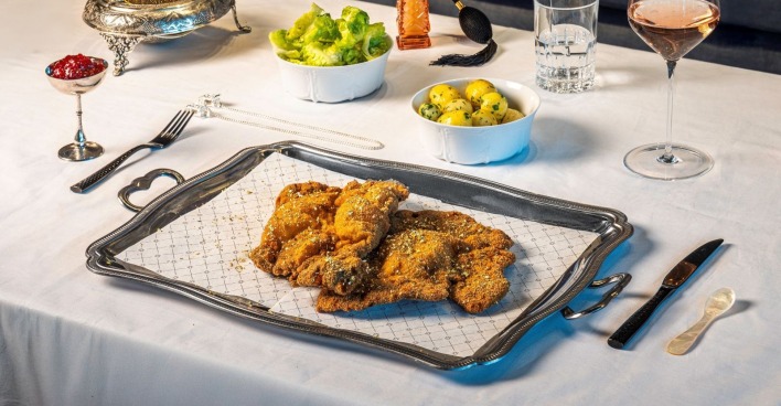 Luxuriös angerichtetes Wagyu-Schnitzel mit Goldflocken auf silberner Platte, dazu Schälchen mit Kaviar, Preiselbeeren, Petersilienkartoffeln und Salat auf weiß gedecktem Tisch.