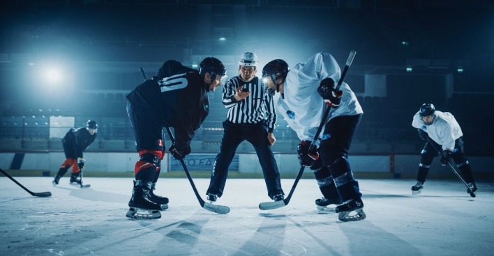 Zwei Eishockeyspieler stehen sich am Eis gegenüber, in der Mitte steht ein Schiedsrichter