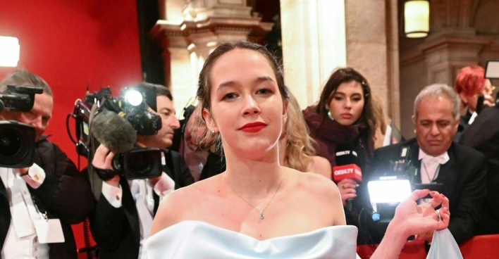 Lena Schilling in einem weißen Kleid beim Wiener Opernball, umgeben von Kameras und Journalisten, während sie mit einer politischen Botschaft protestiert.