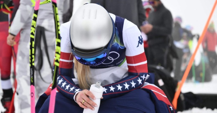 US-Skirennläuferin Lindsey Vonn im Olympia-Rennanzug sitzt erschöpft im Schnee, während das Abfahrtstraining in Cortina wegen Nebel und schwieriger Bedingungen gestoppt ist.