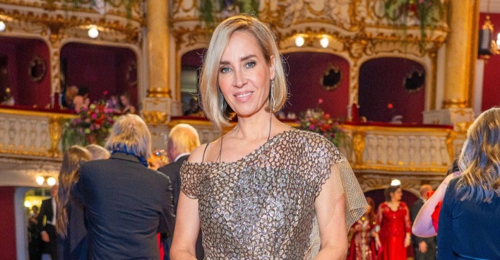 Nadja Bernhard in einem goldfarbenen Abendkleid in einem prunkvollen Theatersaal mit roten Logen im Hintergrund.