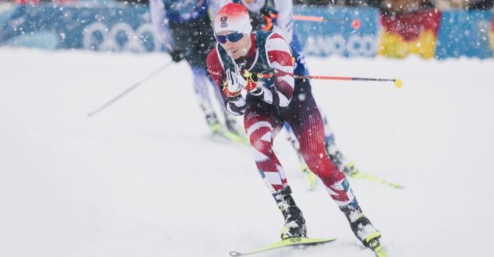Ein österreichischer Nordischer Kombinierer in rot-weißem Rennanzug läuft bei starkem Schneefall in der Loipe während des olympischen Teamsprints in Val di Fiemme.