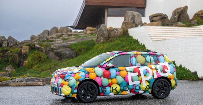 Ein Skoda Epiq steht vor einem grünen Hügel auf dem ein Haus und Felsen zu sehen sind. Das Auto hat keine einheitliche Farbe, vielmehr sind runde "Bälle" in verschiedensten Farben aufgetragen.