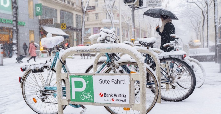 Verschneite Fahrräder an einem Abstellplatz in einer Wiener Einkaufsstraße, während dichter Schneefall und Passanten mit Schirmen das winterliche Stadtbild prägen.