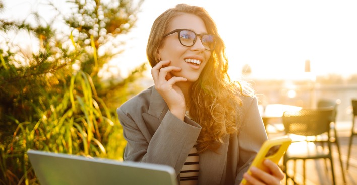 Junge gut gelaunte Frau mit Smartphone in der Hand vor ihrem Notebook | Credit: iStock.com/Maksym Belchenko