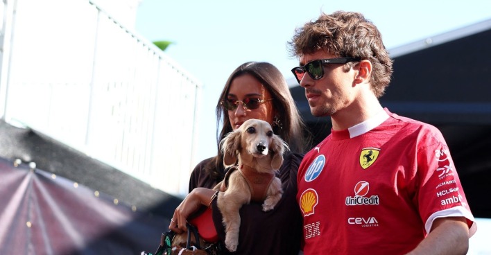 Charles Leclerc in Ferrari-Teamkleidung steht mit Alexandra Saint Mleux und Dackel Leo nebeneinander im Fahrerlager.