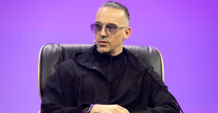 RAF Camora sitzt auf einer Bühne bei einem Interview oder Panel, trägt schwarze Kleidung und eine Sonnenbrille und spricht über seine Rückkehr von Dubai nach Wien nach den Iran-Angriffen.