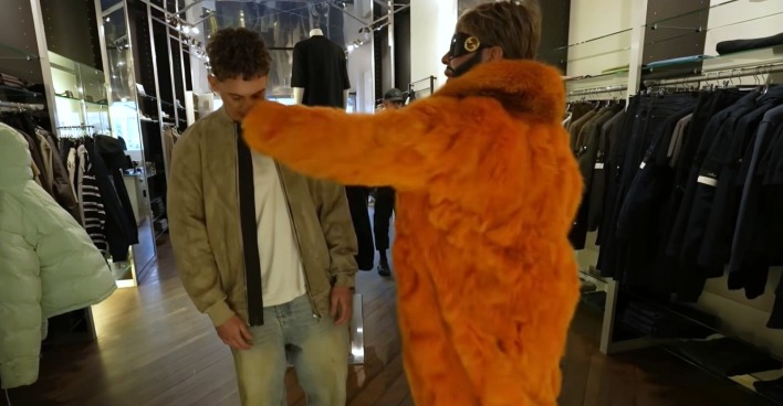 Harald Glööckler in einem auffälligen orangefarbenen Pelzmantel holt in einem Modegeschäft zum Schlag gegen einen jungen Mann aus, während Kleidung an den Wänden hängt.