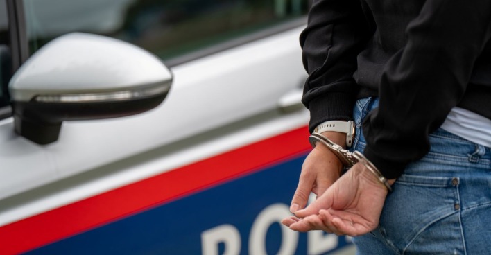 Mann in Handschellen, dahinter steht ein Polizeiauto