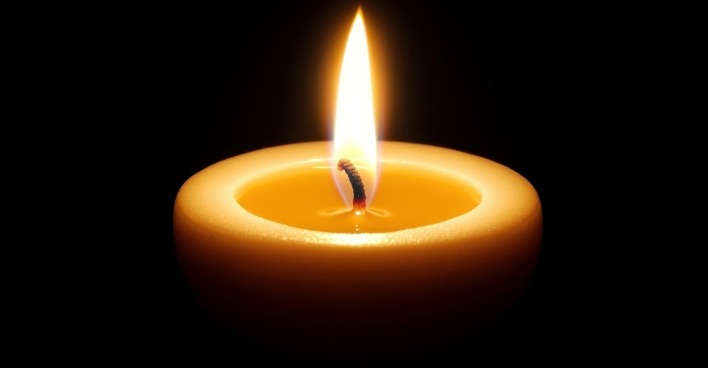 Eine einzelne, helle Kerze sitzt in der Mitte eines schwarzen Hintergrundes, deren warme gelbe Flamme einen sanften Schein auslöst.
