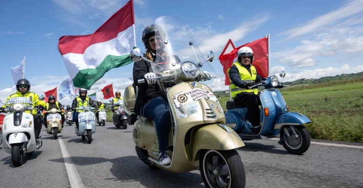 Eine Gruppe von Vespa-Fahrern auf weißen, blauen und goldenen Vespas fahren auf einer Landstraße. Alle tragen einen Helm, manche fahren mit der italienischen Flagge.