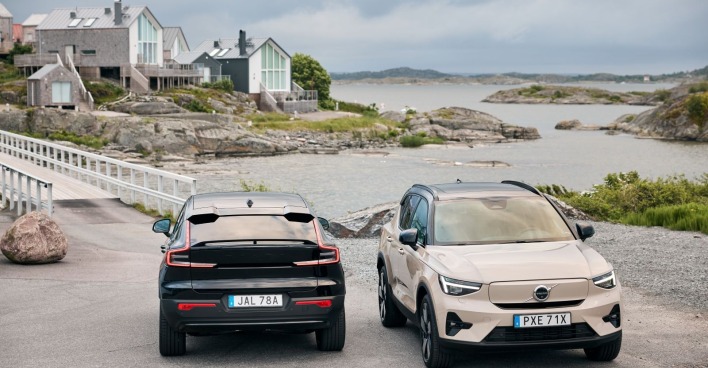 Ein schwarzer Volvo EC40 und ein hellbrauner Volvo EX40 parken am Rande einer Meeresbucht. Im Hintergrund sind einige Häuser zu sehen, am Himmel einige Wolken.