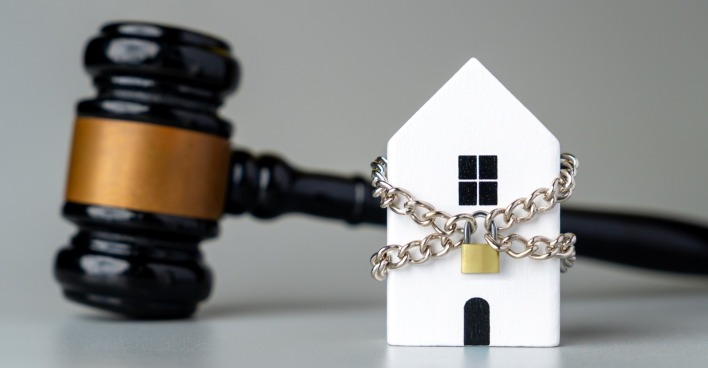 Ein Miniatur-Haus-Modell in einer Metallkette verpackt und ein Vorhängeschloss mit einem Gavel.