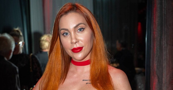 Tara Tabitha posiert mit roten Haaren und auffälligem roten Lippenstift bei einem Event und blickt direkt in die Kamera.